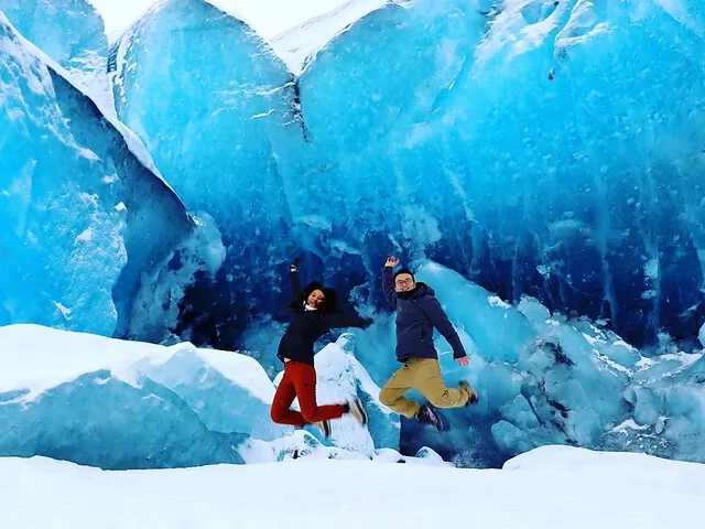 Two people jumping excitedly in front of a massive blue ice formation.