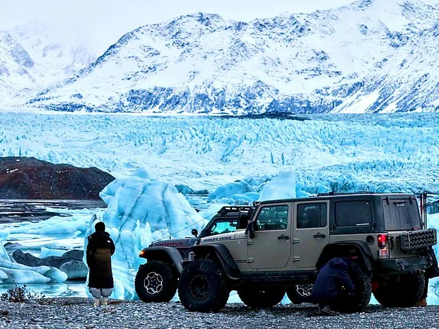 Two people standing near a Jeep parked in front of a massive glacier.