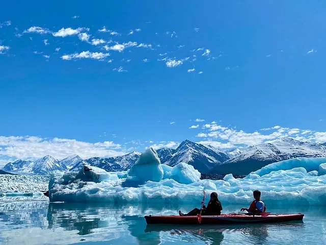 Two people kayaking in front of a large glacier under a blue sky