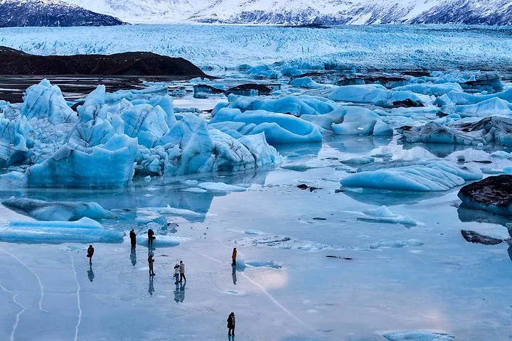 Several people walking on a frozen glacial lake near large blue ice formations.