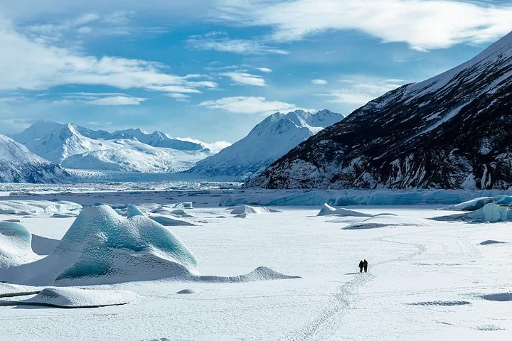 Two distant figures walking on a vast snow-covered glacier surrounded by mountains.