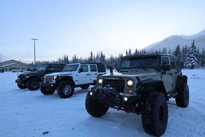 Three off-road vehicles parked on a snowy field near a building and mountains.