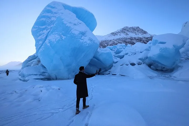 A person taking a picture of large chunks of blue ice on a snowy surface.
