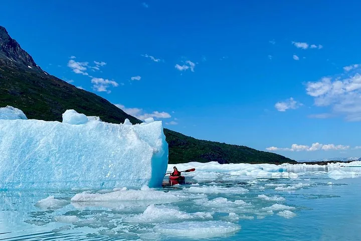 Person in a red kayak paddling past a massive blue iceberg