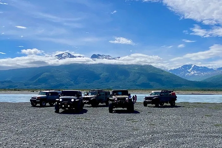 Group of off-road vehicles parked on a rocky riverbank with mountains behind