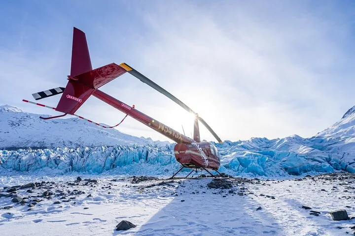 Red helicopter landed on a snowy landscape with blue glacial ice nearby.