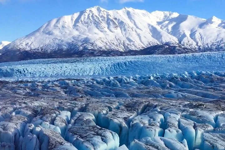 Expansive view of a cracked blue glacier with snow-capped mountains behind.