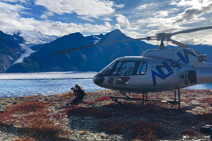 Photographer kneeling near a helicopter with Knik Glacier and mountains in background.